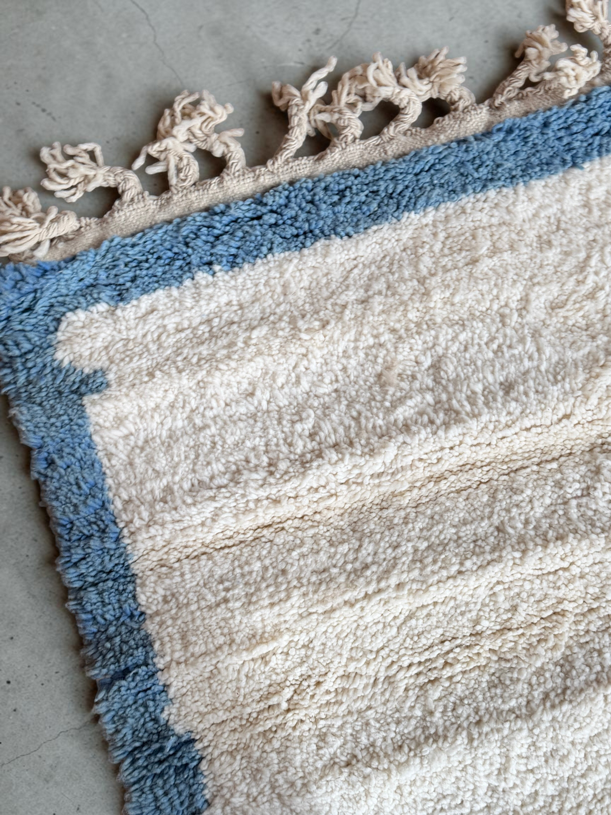 Tapis YAAS Alya en laine naturelle noué à la main avec bordure bleue et franges décoratives sur sol en bois